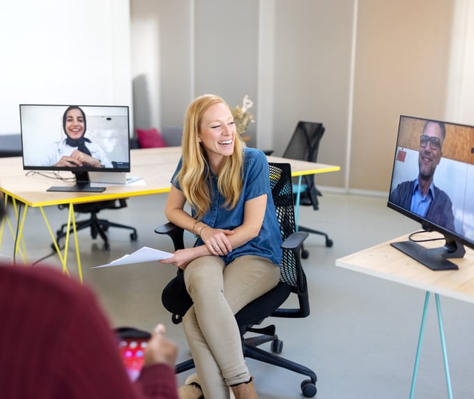 Alt text: Woman seated in an office smiling during a hybrid meeting, with two coworkers appearing on desktop monitors for a video call and another person blurred in the foreground.