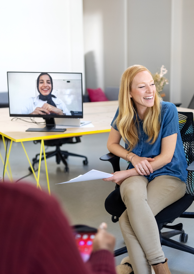 Alt text: Woman seated in an office smiling during a hybrid meeting, with two coworkers appearing on desktop monitors for a video call and another person blurred in the foreground.