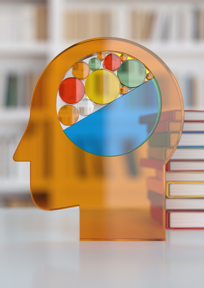 Transparent orange silhouette of a human head with a colorful abstract brain made of circles and geometric shapes, set on a table beside stacks of books with blurred bookshelves in the background.