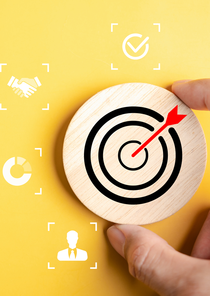 Hand placing a round wooden token with a bullseye and red arrow on a yellow background, surrounded by white business icons representing goals, partnership, analytics, settings, performance, and leadership.